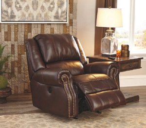 The Chestnut Collinsville leather recliner with a nailhead trim. next to a brown wooden side table with a lamp on it.