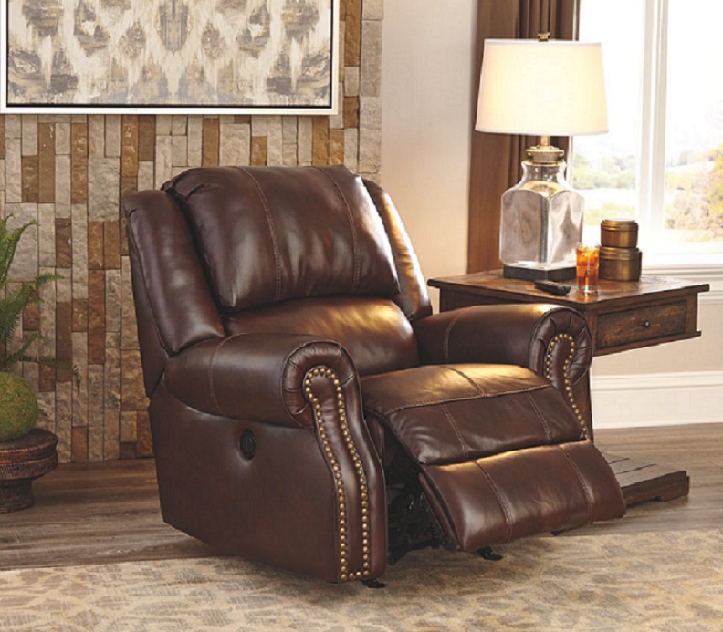 The Chestnut Collinsville leather recliner with a nailhead trim. next to a brown wooden side table with a lamp on it.