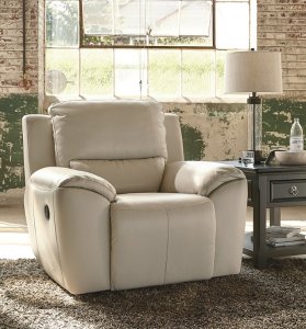 The white Valeton leather recliner chair on top of a gray fluffy rug with a dark brown side table next to it. On the side table is a class lamp and a drawer for storage.