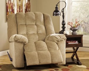 The sand Colored Ludden recliner with lots of plush next to a side table with storage space. A lamp is on top of the side table and there is abstract wall are behind the recliner.
