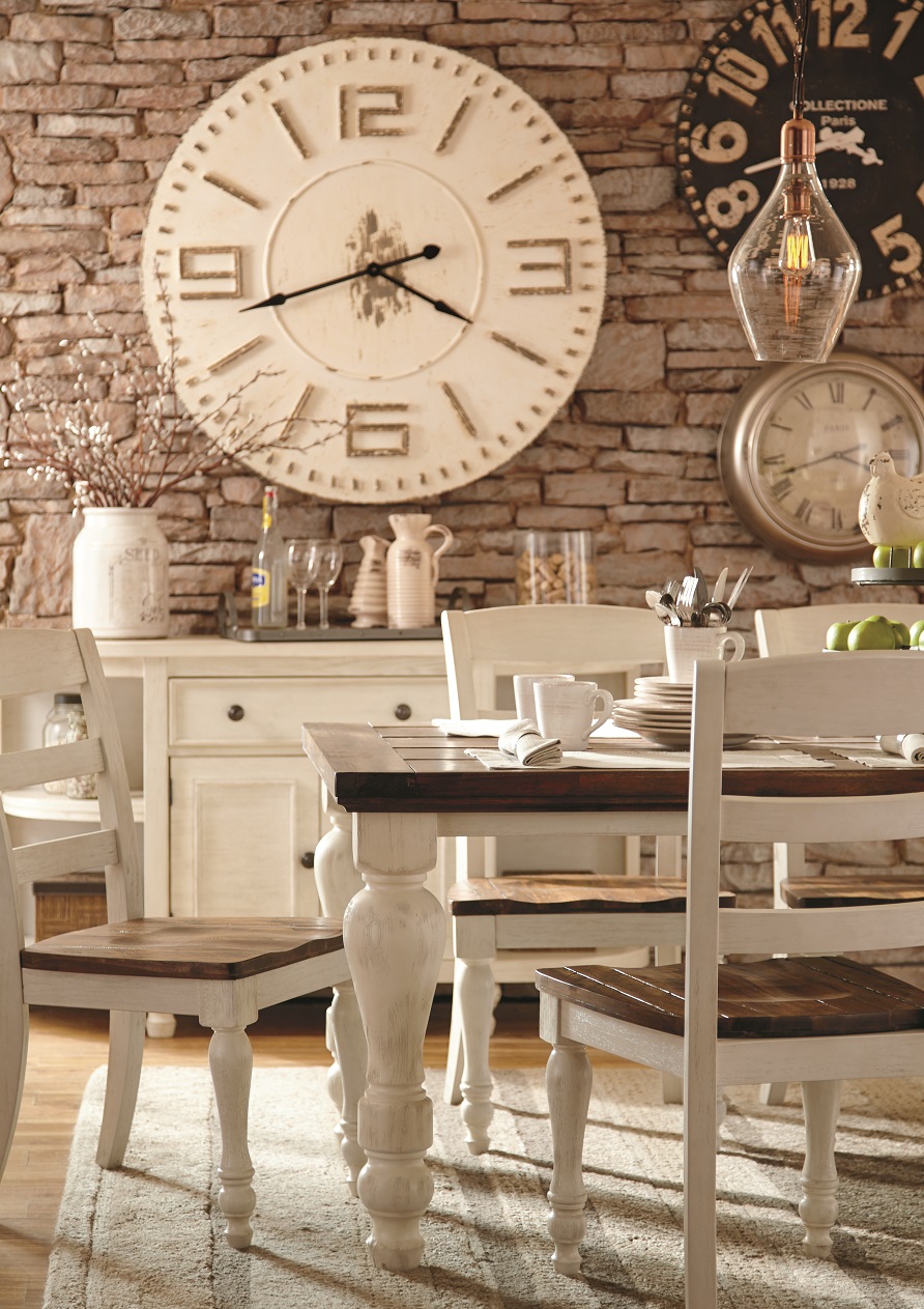 A dining room table in front of a server with a large clock on a brick wall.