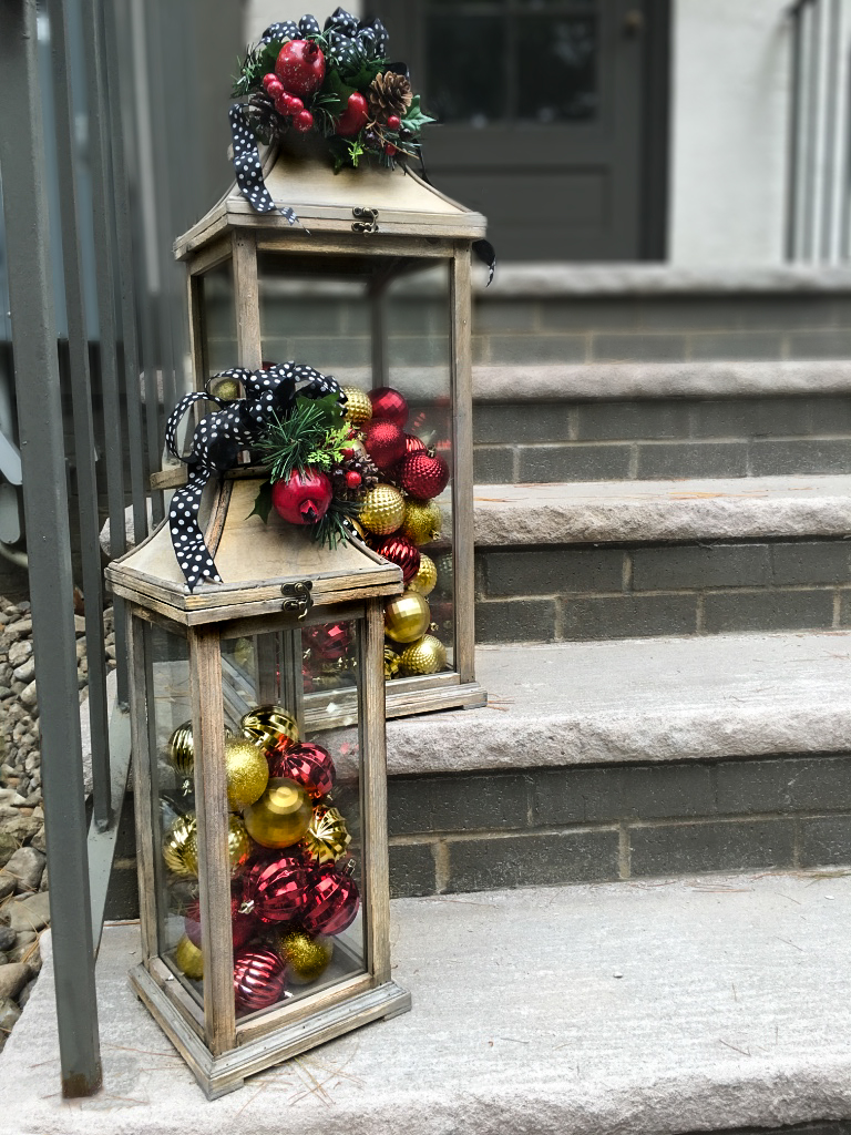 Glass lanterns used as holiday decorations that are filled with Christmas tree ornaments, decorated with ribbon, and Christmas garland.