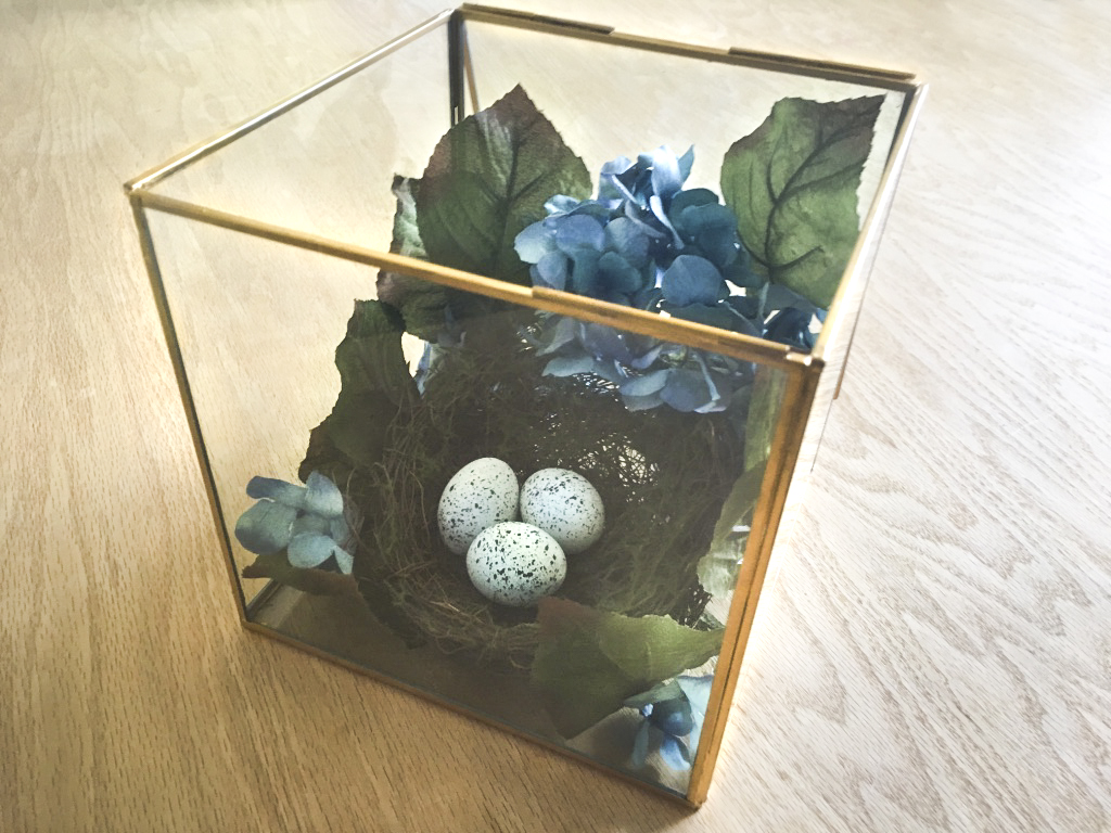 Glass box filled with faux flowers, eggs, nest, and faux eggs for a spring home decoration.