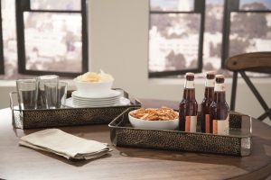 antique bronze tone metal serving trays with drinks and snacks on a coffee table with two windows in the background.