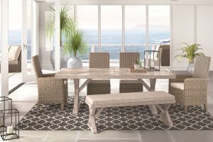 Outdoor dining room table with matching chairs. The dining room is overlooking the ocean with several indoor plants in the background. On the table is a tray with a succulent and a candle holder with a candle inside. Underneath the dining room table is a black patterned rug.