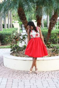 Model with red skirt and floral top standing on cobblestones in a park with palm trees behind her.