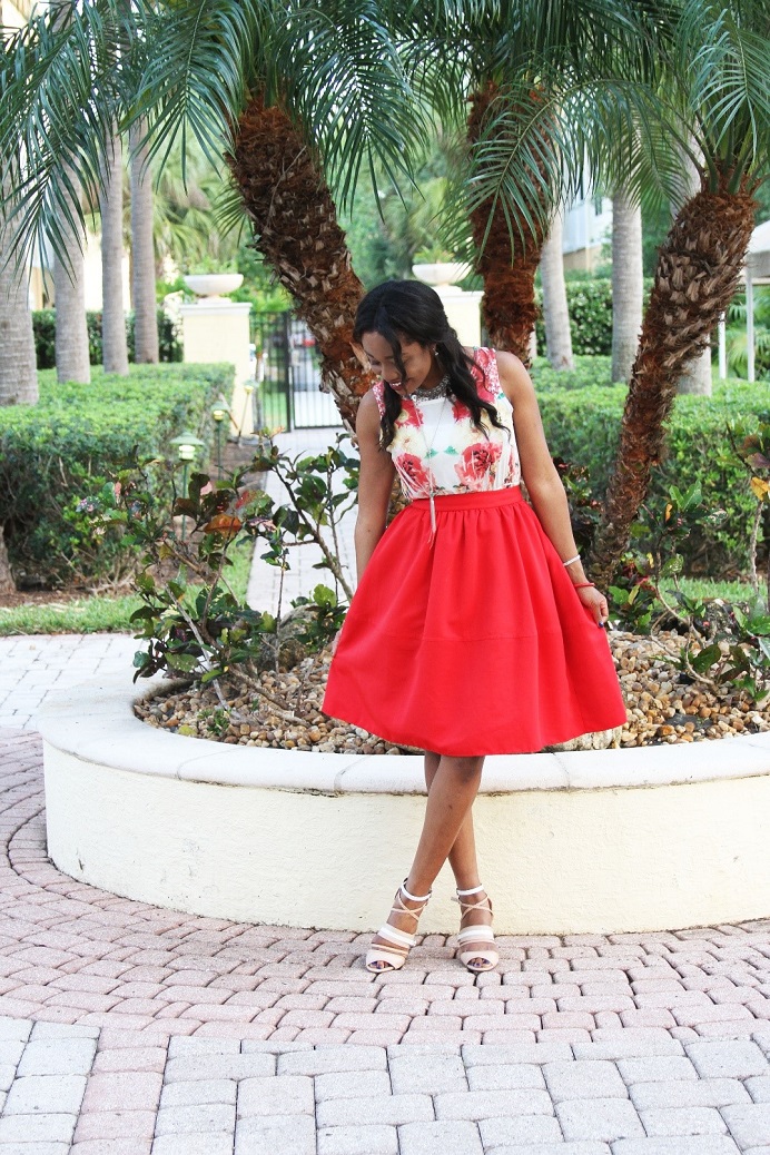 Model with red skirt and floral top standing on cobblestones in a park with palm trees behind her. 