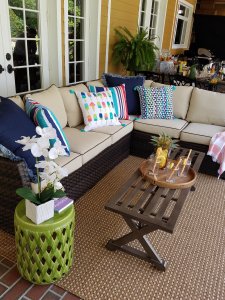 An outdoor cushioned sectional with bright and vibrant fun pillows on top. An outdoor table with a tray of lemonade on top with a small neon green side table with greenery for decoration.