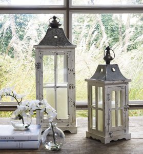 Set of two white washed candle lanterns on a table with with a window behind them overlooking ferns.