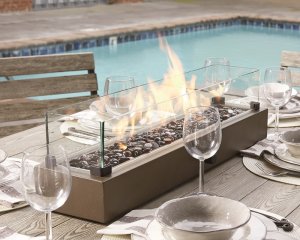 a sleek rectangular tabletop fire bowl in the center of a set table with a pool in the background.