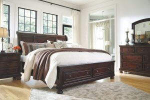 Brown wooden contemporary bed in a white room with windows behind the bed and a lush comforter.