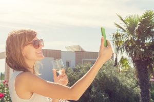 Smiley girl using cellphone and drinking wine outdoors.