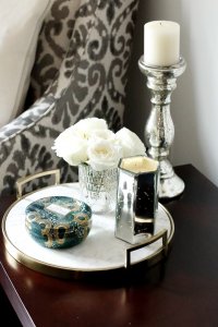 A flat marble tray with a vase of flowers and a candle on top with a accent chair in the background.