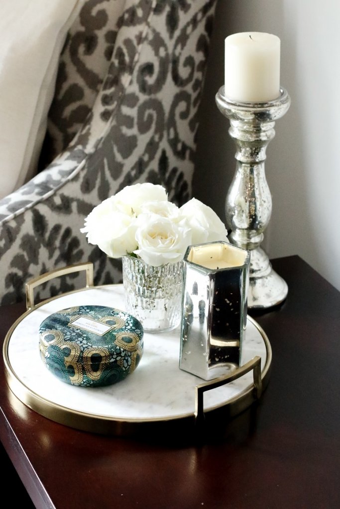 A flat marble tray with a vase of flowers and a candle on top with a accent chair in the background. 