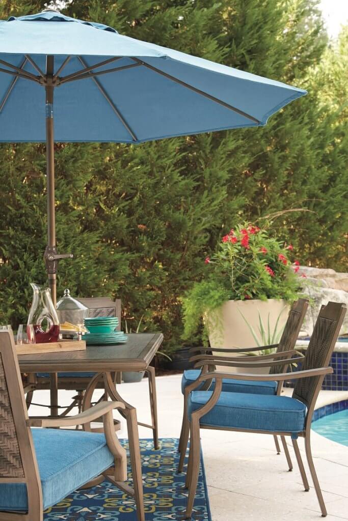 An outdoor dining table on a porch with a blue umbrella hovering above the table. 